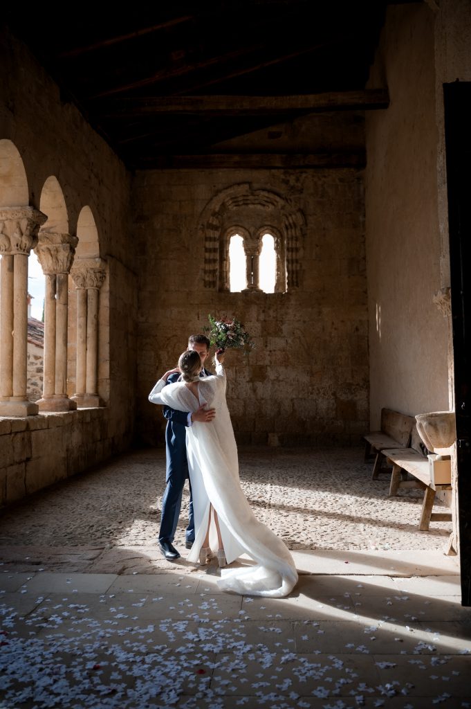 Novios abrazados en el claustro románico de Sotosalvos, Segovia, rodeados de arcos y columnas de piedra, con confeti blanco en el suelo y luz natural lateral entrando por los arcos.